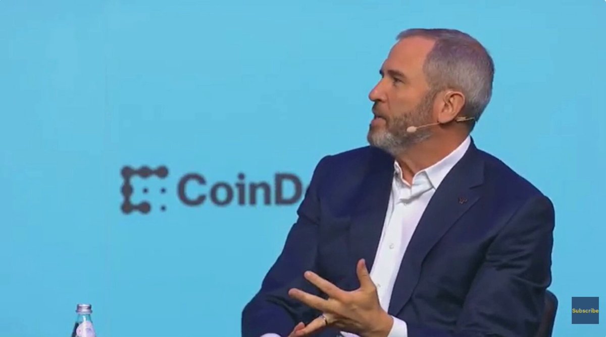 The image features a man wearing a suit, sitting at a table with his hands together while talking to someone. The background of the image shows a blue wall with the word "CoinD" written on it. There are also two bottles visible in the scene - one near the left edge and another towards the right side.