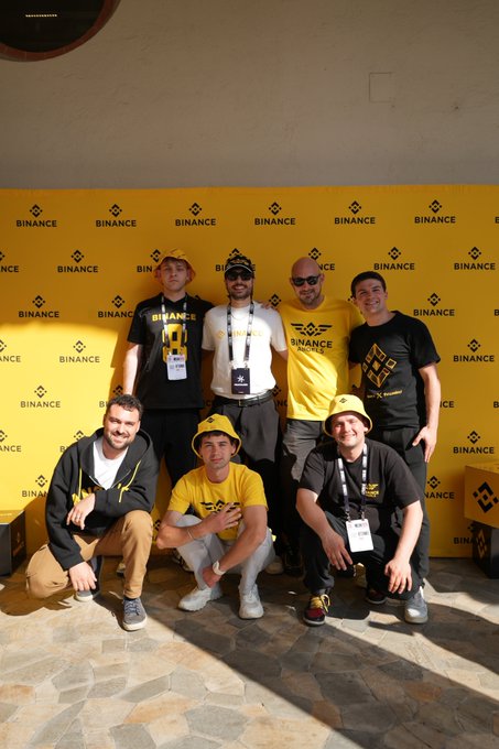 The image features a group of six men posing for a photo against a yellow backdrop that says "Binance" on it. The men are wearing matching yellow shirts, which suggests they might be part of an event or gathering related to Binance. They are standing in front of a wall with the word "ETHMILAN" written on it, indicating that this meetup took place at a location in Milan, Italy. The group appears to be enjoying themselves and is likely proud to represent Binance during their time together.