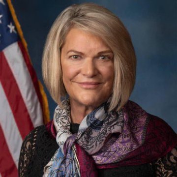 A professional portrait of Senator Cynthia Lummis (Republican of Wyoming) smiling with blonde hair, wearing a black top with a colorful patterned scarf, posed beside an American flag. The image accompanies news about her confidence that the Senate will vote on the Clarity Act before the August recess.