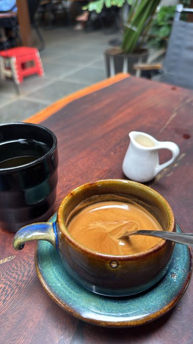 A ceramic cup of coffee with latte art sitting on a saucer on a wooden table, accompanied by a small white pitcher of milk and a dark mug in the background.