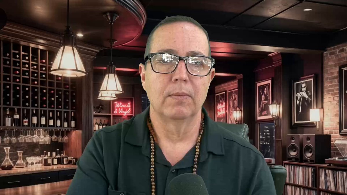 The image features a man wearing glasses sitting at a bar, with a microphone in front of him. The background includes several wine bottles displayed behind him. There are also some posters hanging on the wall above the bar. The caption "Why I am NOT SElling my XRP? youtu.be/8W50V7spdUw" is written below the man, which seems to be related to a post about selling XRP.