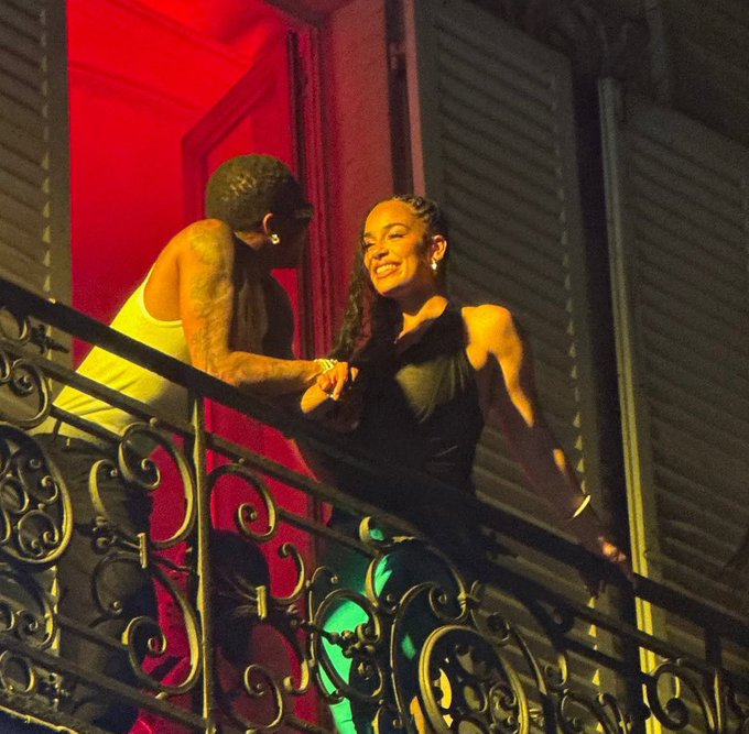 A nighttime photo of a man in a white tank top and a woman in a black halter top smiling at each other on an ornate wrought-iron balcony with red ambient lighting.