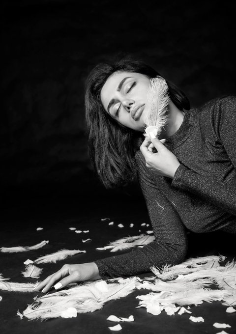 A black and white photograph of a woman lying down, peacefully holding a white feather to her face amidst scattered feathers and rose petals on a dark surface.