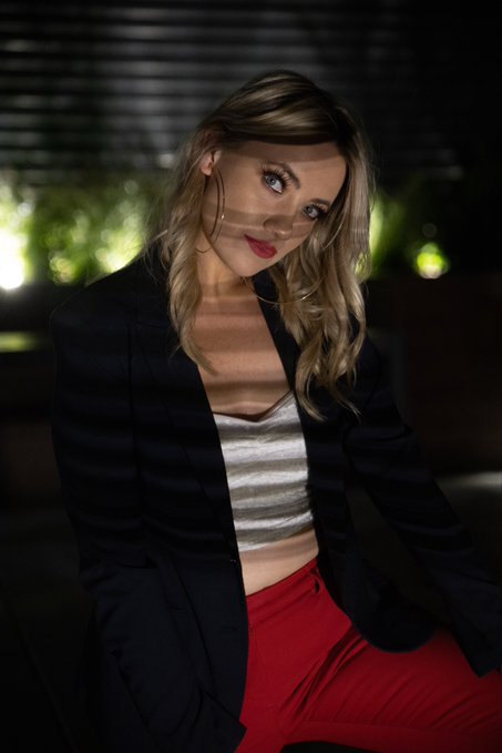 A blonde woman with a stylish haircut poses indoors, wearing a black blazer, white top, and red pants, with light filtering through window blinds casting striped shadows across her.