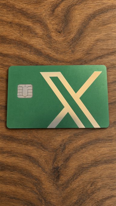 A green card with a gold geometric 'X' design and an EMV chip rests on a wooden surface.