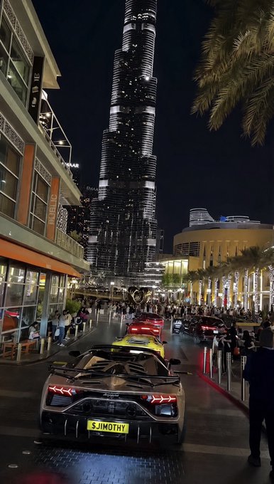 A nighttime street scene in Dubai featuring the Burj Khalifa, luxury cars including a Lamborghini with the license plate 'SJIMOTHY', and crowds of people.