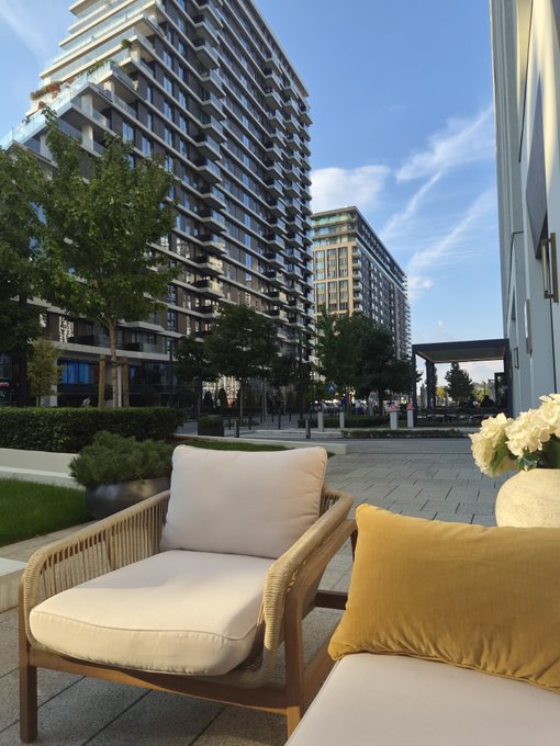 An outdoor patio area features a wicker chair with white cushions and a yellow pillow, set against the backdrop of a tall modern apartment complex under a blue sky.
