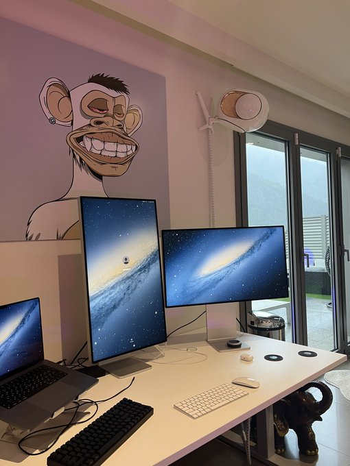 A home office workspace features dual monitors and a laptop displaying galaxy wallpapers, set against a wall adorned with a Bored Ape NFT artwork—tying directly into the crypto/NFT "gApE frens" theme of the post.