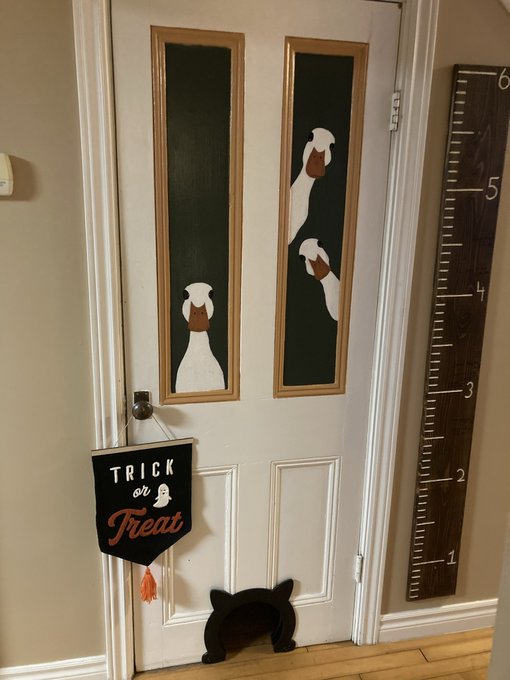 A white door features painted dog artwork in its window panels, a 'Trick or Treat' banner hanging from the handle, and a black cat-shaped cutout at the base, with a wooden height chart mounted on the adjacent wall.