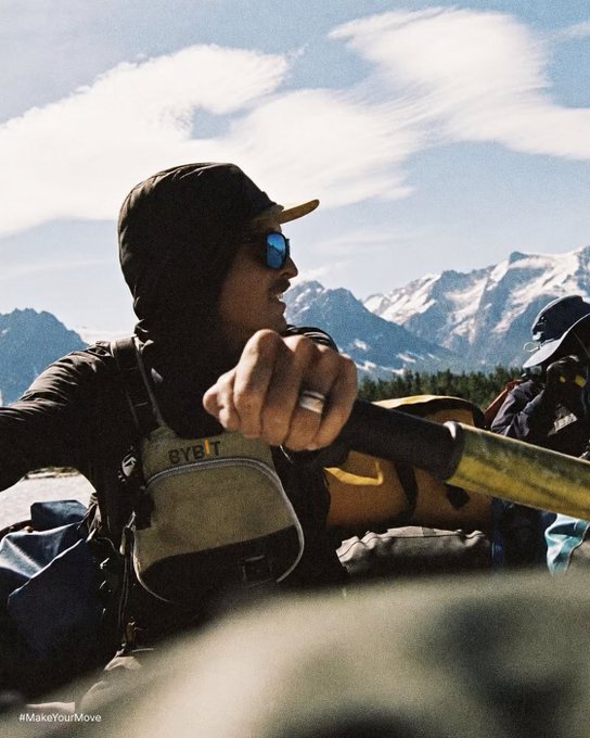A man wearing sunglasses, a cap, and a hooded shirt, sitting outdoors with snow-capped mountains in the background; he is extending his fist toward the camera and has '#MakeYourMove' written in the bottom corner.