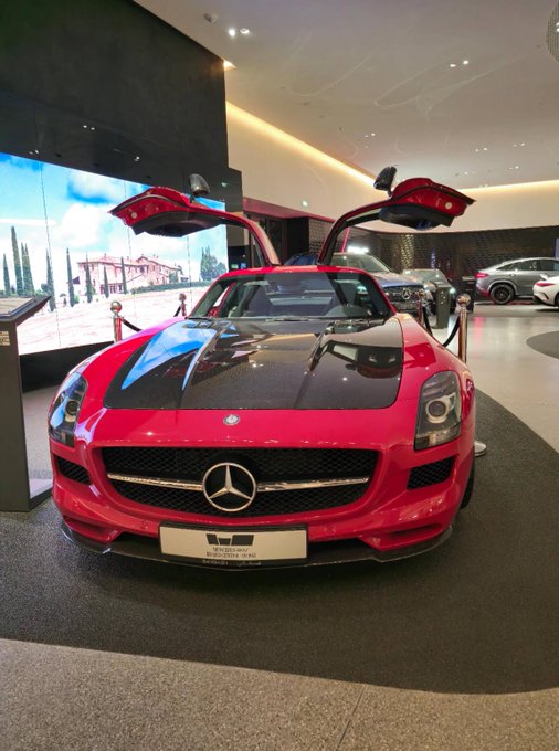 A red Mercedes-Benz SLS AMG with gullwing doors open is displayed in a showroom, prominently featuring the AMG logo on its grille. The luxurious sports car image contrasts with the post's message about independent research and crypto/prediction market trading, emphasizing the "don't blindly follow others" theme through its bold, standout presentation.