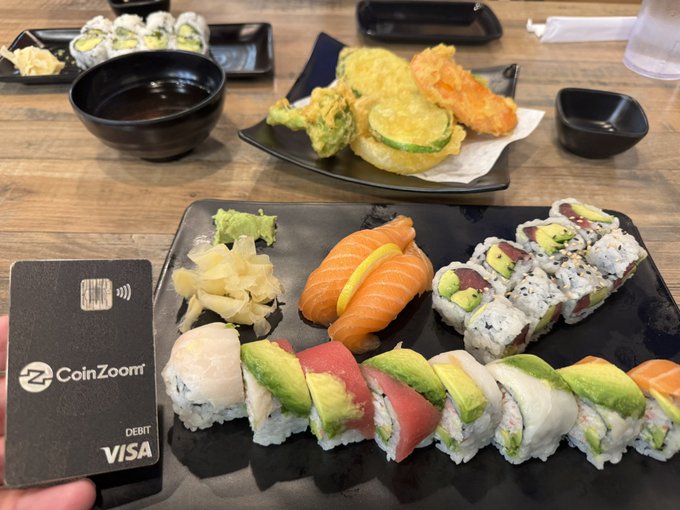 A spread of sushi rolls, nigiri, and tempura is arranged on black plates alongside a CoinZoom Visa debit card and a bowl of miso soup.