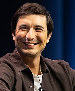 A smiling Asian man with short dark hair wearing a black button-up shirt over a white t-shirt is photographed against a dark blue background.