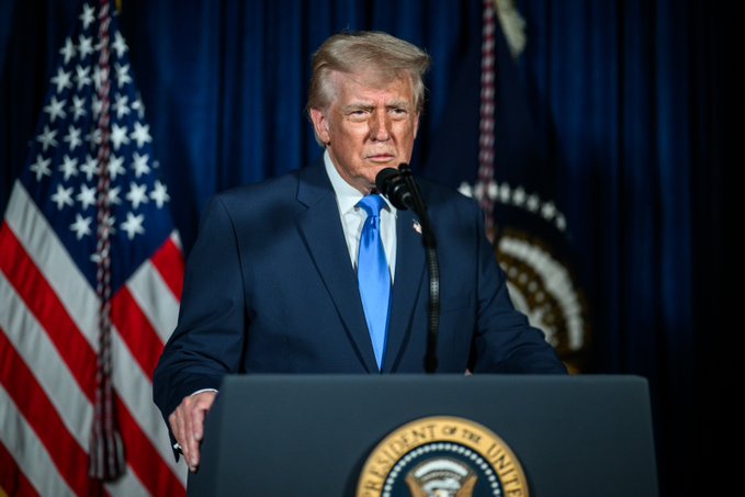 The user wants a concise caption for the image.
I need to identify the key elements:
- **Subject:** Donald Trump.
- **Action:** Standing at a podium, speaking (or about to speak).
- **Setting:** Formal setting, likely the White House.
- **Key Visuals:** US Flag, Presidential Seal on the podium, Blue curtain background.
- **Text:** The seal says "PRESIDENT OF THE UNITED STATES".

The prompt asks for a 1-2 sentence caption describing what is in the image, including key text.

**Drafting the caption:**
- "Donald Trump stands at a podium bearing the Presidential Seal, with an American flag visible behind him."
- "Donald Trump is pictured speaking at a podium featuring the 'President of the United States' seal, with the US flag in the background."

Let's refine it to be concise and accurate.
"Donald Trump stands at a podium with the 'President of the United States' seal, speaking into