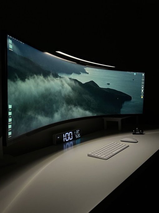 A large curved ultrawide monitor displays a mountainous landscape wallpaper on a sleek desk setup featuring a digital clock showing '1:00', a keyboard, and a mouse.