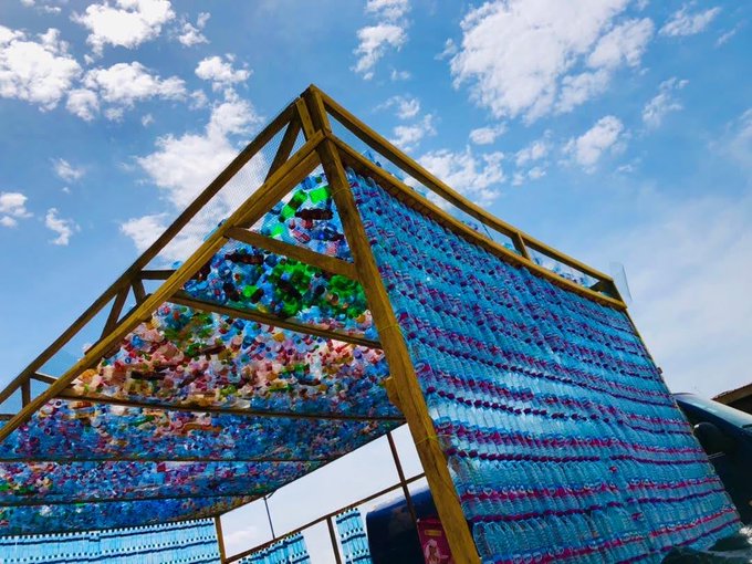 A large outdoor art installation constructed from a wooden A-frame structure filled with rows of blue and multicolored plastic bottles, set against a partly cloudy sky.