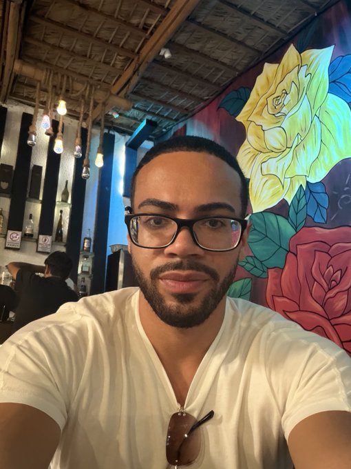 A selfie of a bearded man wearing glasses and a white shirt, taken inside a restaurant or bar with colorful rose mural artwork on the wall behind him.