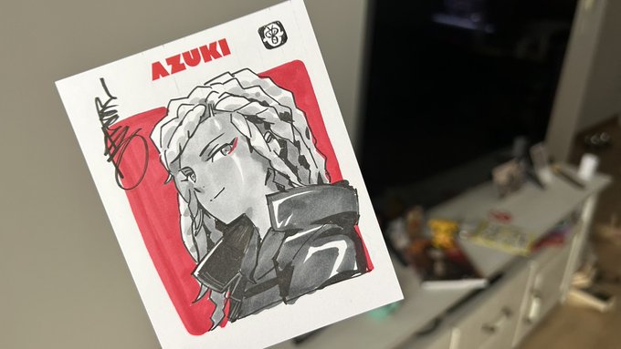 A signed card or print labeled 'AZUKI' featuring an anime-style illustration of a character with long light hair and red eyes against a red background, held up in front of a desk.