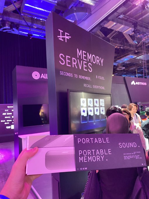 A first-person view at an event holding promotional cards that read 'PORTABLE SOUND. PORTABLE MEMORY.' in front of a 'MEMORY SERVES' booth display.