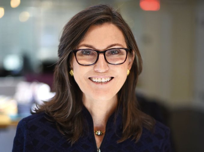 A smiling woman with dark brown hair, wearing tortoiseshell glasses and a navy blue textured jacket, appears in a professional portrait setting. The image likely depicts Cathie Wood, the CEO of ARK Invest, in connection with her recent $21 million purchase of Amazon shares amid growing investment in AI-benefiting companies.