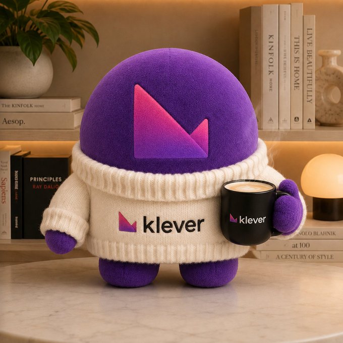 A purple plush mascot character wearing a white sweater and holding a mug, both branded with the 'klever' logo, sitting on a table surrounded by books.