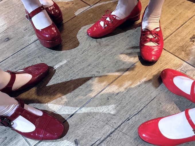 Four pairs of feet in red shoes—including Mary Janes, lace-up flats, and T-straps—form a circle on a rustic wooden floor, perfectly capturing a "coming right up" moment as everyone steps into the frame. The coordinated footwear and arrangement suggest a playful gathering or dance about to begin.