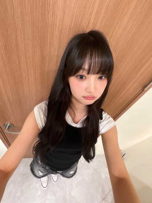 A young woman with long dark hair, wearing a white t-shirt over a black top and ripped jeans, poses for a selfie against a wooden door.