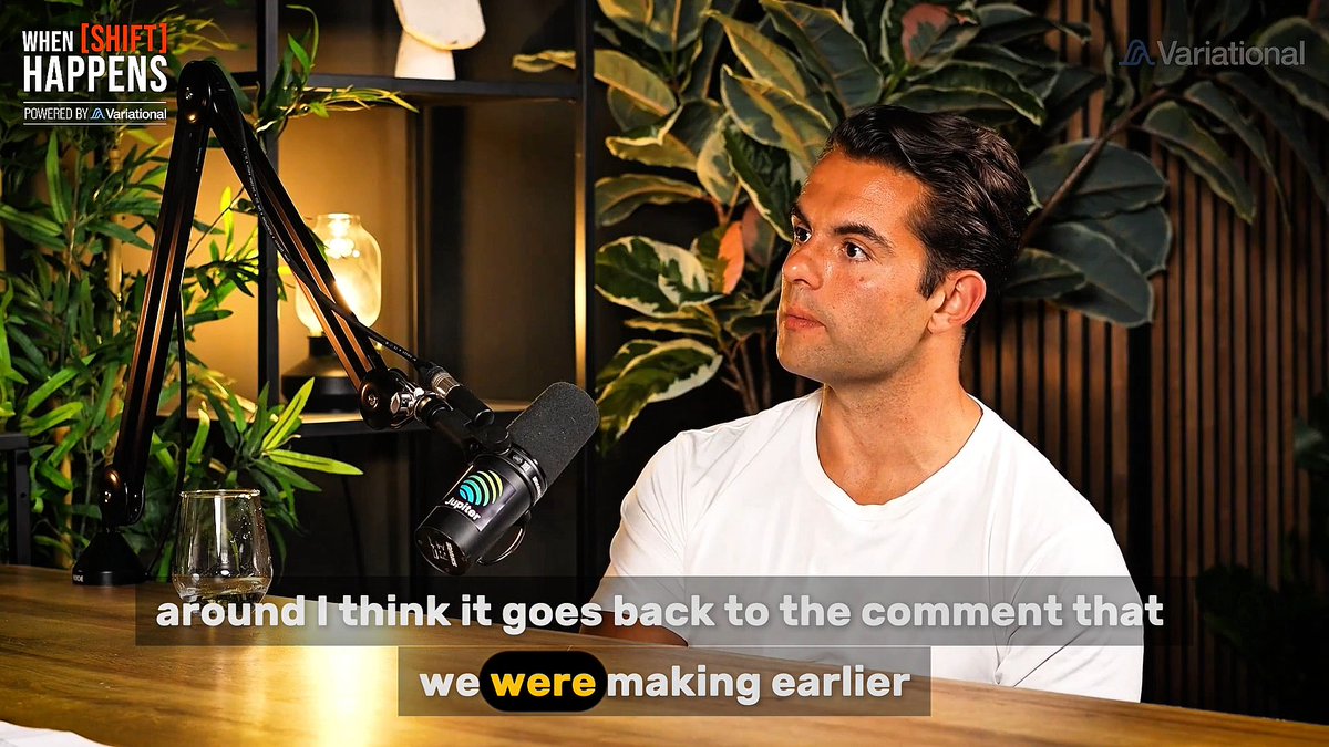 A man in a white t-shirt speaks into a podcast microphone on the show 'When [SHIFT] Happens,' powered by Variational, with subtitles reading 'around I think it goes back to the comment that we were making earlier.'