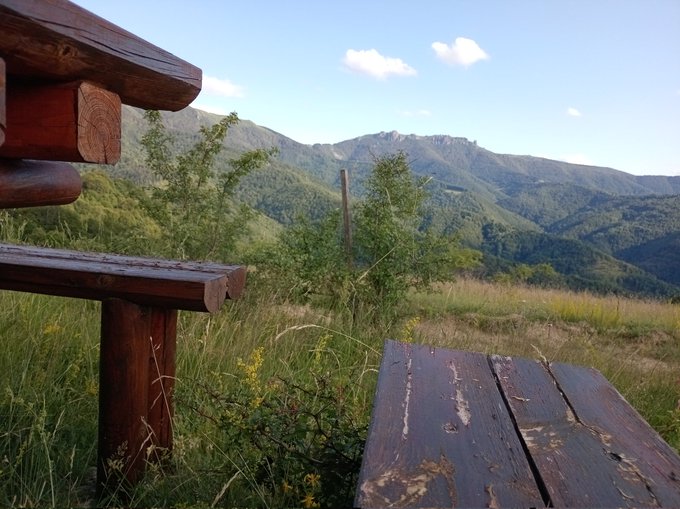A scenic mountain view from a wooden shelter or picnic area, with weathered wooden planks in the foreground and rolling forested hills under a partly cloudy blue sky in the background.