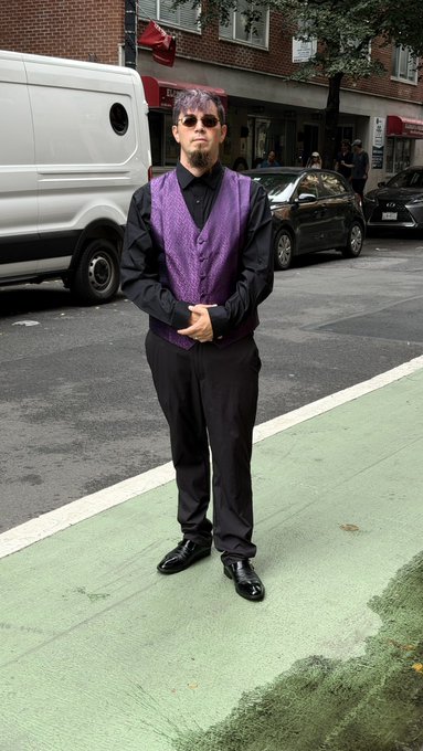 A man with purple hair, sunglasses, a goatee, and a purple patterned vest over a black shirt stands on a green bike lane with a white van parked on the city street behind him.