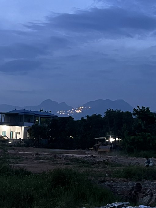 A twilight landscape photo showing a building with lit windows in the foreground, silhouetted mountains, and scattered city lights visible in the valley beyond.