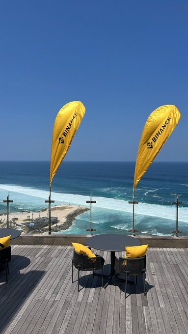 Two yellow Binance branded flags stand on a wooden deck overlooking a scenic beach with turquoise ocean waves and rocky shoreline under a clear blue sky.