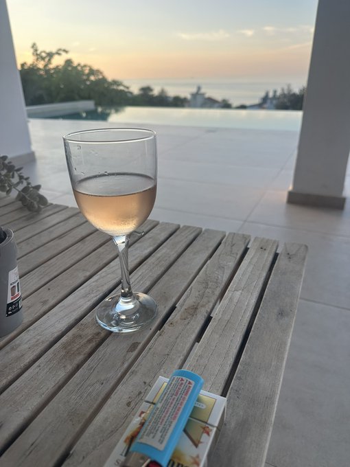 A glass of rosé wine sitting on a wooden outdoor table overlooking an infinity pool and ocean at sunset, with a can and cigarette pack nearby.