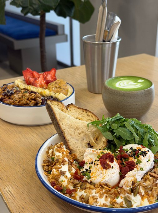 A cafe meal featuring a bowl with poached eggs, grains, toasted bread, arugula, and seeds, accompanied by a matcha latte in a ceramic cup, with a fruit and granola bowl in the background.