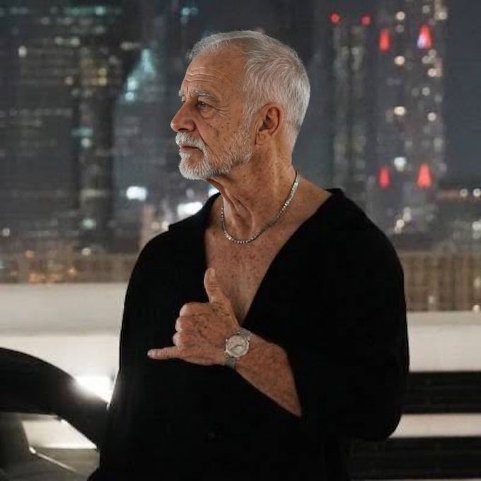 An older man with gray hair and a white beard, wearing a black v-neck shirt and a silver chain necklace, gestures with his hand against a nighttime city skyline backdrop.