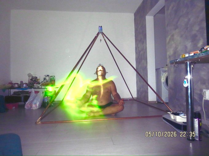 A shirtless man meditates in a lotus position inside a copper pyramid frame, with a date stamp reading '05/10/2026 22:35' and glowing green light effects in a sparsely furnished room.