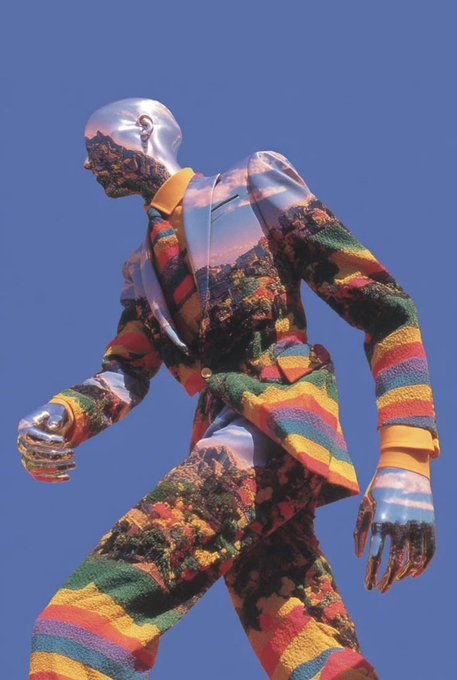 A mannequin wearing a colorful patterned suit featuring landscape imagery on the jacket and rainbow stripes on the sleeves and pants, set against a blue sky.