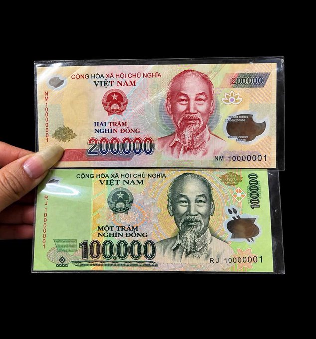 A hand holds two Vietnamese banknotes featuring Ho Chi Minh: a 200,000 Dong note and a 100,000 Dong note.
