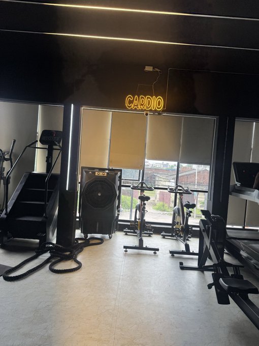 An interior view of a gym with cardio equipment, featuring a neon 'CARDIO' sign on a dark wall and large windows looking out onto a cityscape.