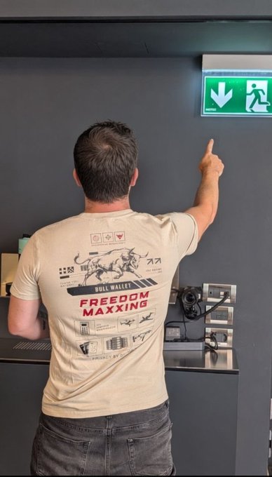A person points towards a green "Exit" sign while wearing a t-shirt that says "BULL WALLET FREEDOM MAXXING", illustrating the need for emergency exits from centralized systems.