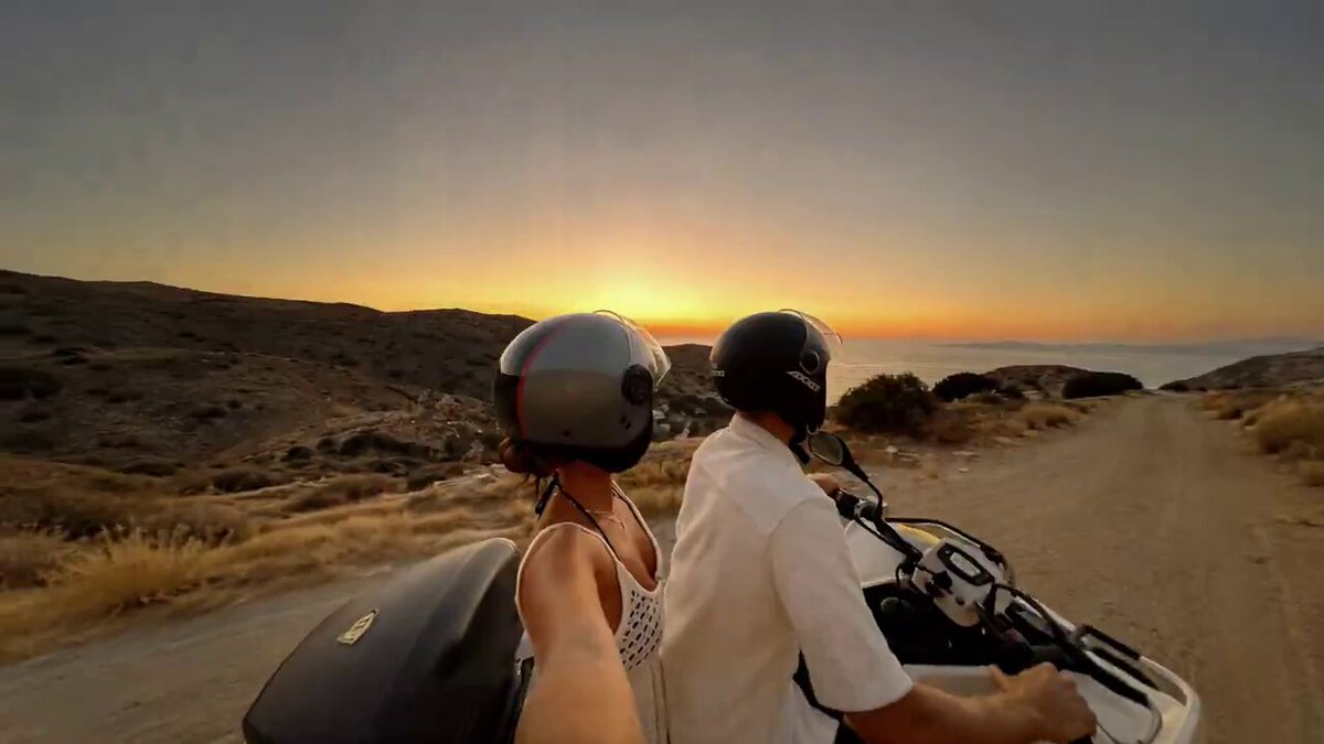 The image shows two people riding motorcycles, with one person wearing a helmet and the other not. They are both looking at their phones while riding down a dirt road. The background features a beautiful sunset over a desert landscape.