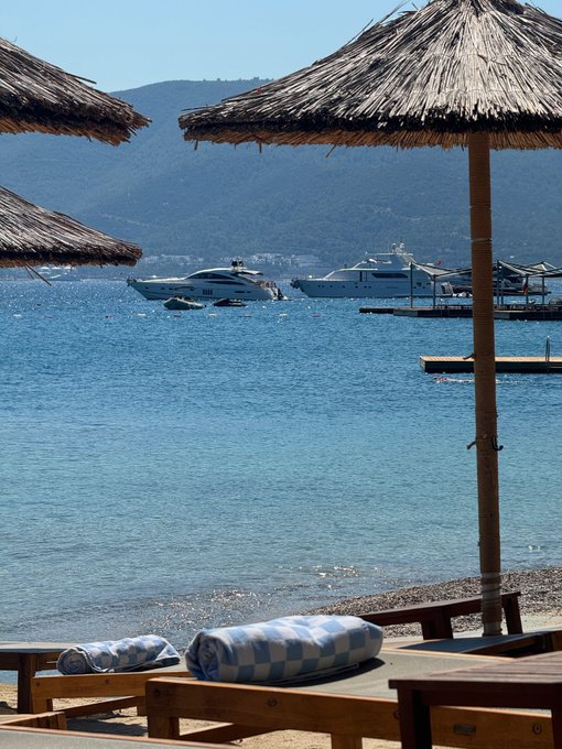 A sun-soaked beach scene features thatched parasols and blue-and-white checkered towels rolled up on wooden lounge chairs, with two luxury yachts anchored in the sparkling turquoise bay beyond. The image captures the "enjoy life" mantra, evoking the Mediterranean leisure that successful bitcoin investors can afford during market downturns.