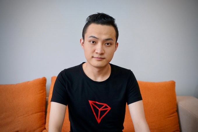 A man wearing a black t-shirt with a red triangle logo sitting on a couch with orange pillows.