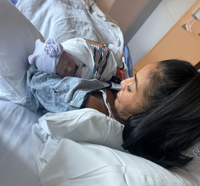 A woman lies in a hospital bed holding a newborn baby wearing a striped beanie hat, with hospital bedding visible around them.