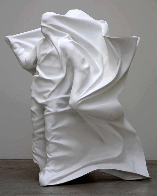 A white sculpture depicting a figure completely draped in flowing fabric, creating dynamic folds and curves on a concrete floor against a gray wall.