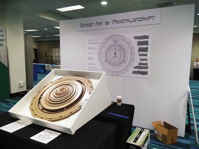 A convention booth displaying Bitcoin Art by FractalEncrypt, featuring a large spiral Bitcoin-themed artwork in a white frame alongside an informational poster.