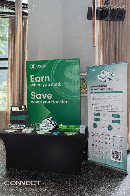 A USDD promotional booth at a Singapore event in October 2026 with banners reading 'Earn when you hold. Save when you transfer.' and a display of branded merchandise.