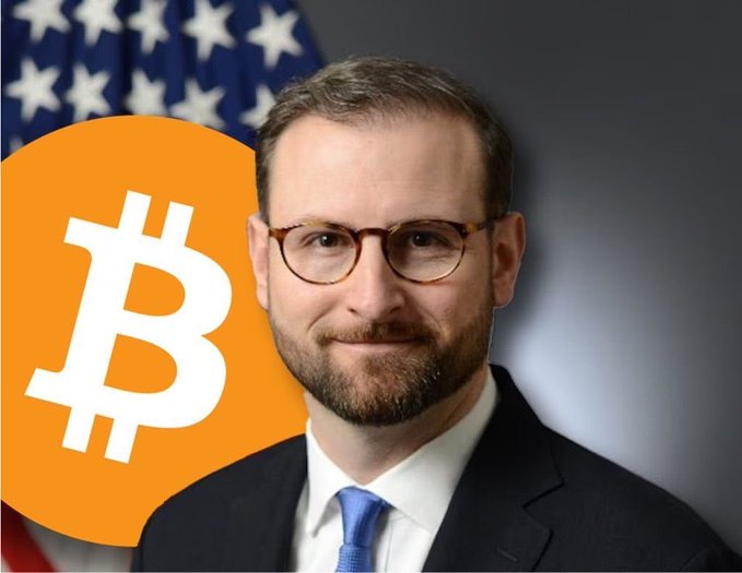 A man in a suit and glasses stands in front of an American flag, with an orange Bitcoin logo overlaid on the left side of the image, visually connecting U.S. policy discussions to cryptocurrency amid calls for regulatory clarity on digital assets.