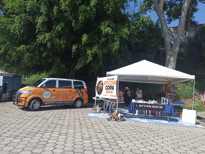 An outdoor Bitcoin-themed promotional setup featuring an orange "Bitcoin Copa" van parked next to a white tent with "Bitcoin Berlin" branded table.