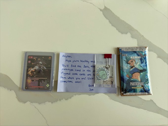 Trading cards and a handwritten note on a marble surface; the note is addressed to "Akylles" and mentions finding a "Zero" prototype card, accompanied by sealed Azuki participant cards.